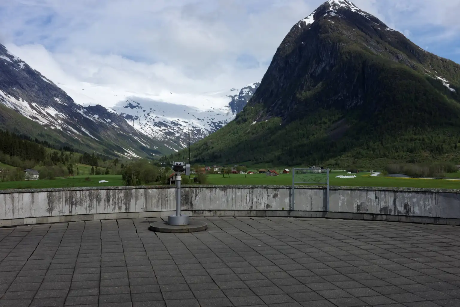 Bilde fra utkikksposten ved det norske isbremuseet. Høye, snødekte fjell kan sees i horisonten. I forgrunnen er det bilde av en kikkert på utkikksposten.
