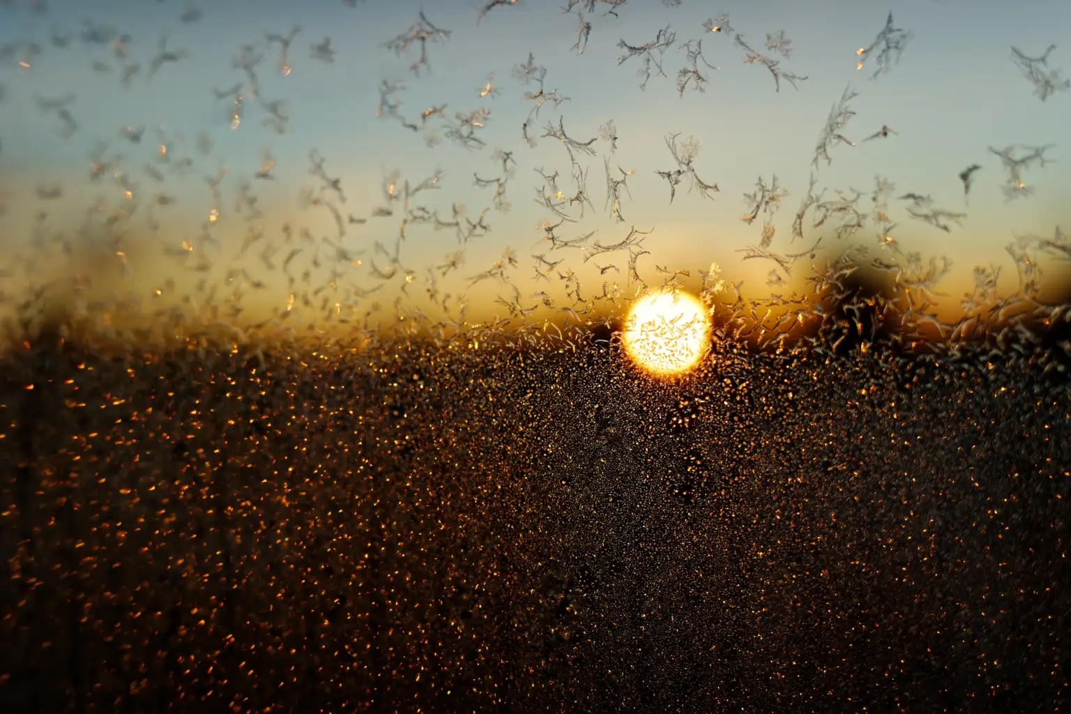 Nærbilde av frostkrystaller på et vindu med soloppgangen i bakgrunnen. Bildet er farget oransje av sollyset.