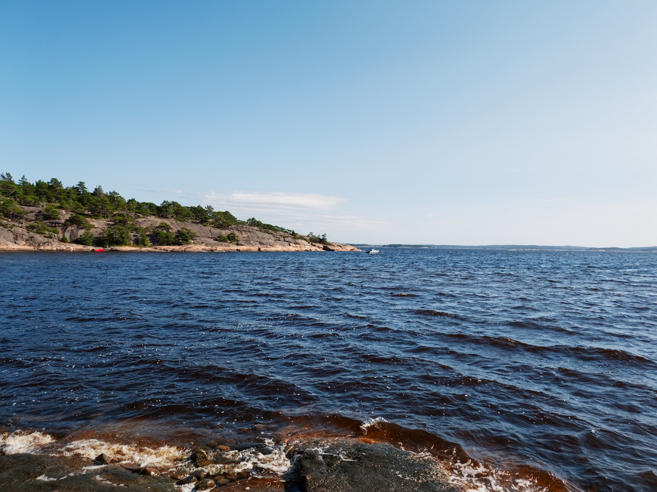 Bilde tatt ved en strand med utsikt over sjøen. Himmelen er blå, og små bølger i det klare vannet slår mot stranden. I horisonten ligger en liten båt og duver ved en holme.